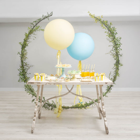 Mesa Candy diseñada y creada por By Ana Laborda para una Comunión. Mesa en tono piedra claro, con dos globos grandes detrás en medio, en colores ácidos, amarillo y azul, flanqueada por dos semiarcos cubiertos de enredadera verde